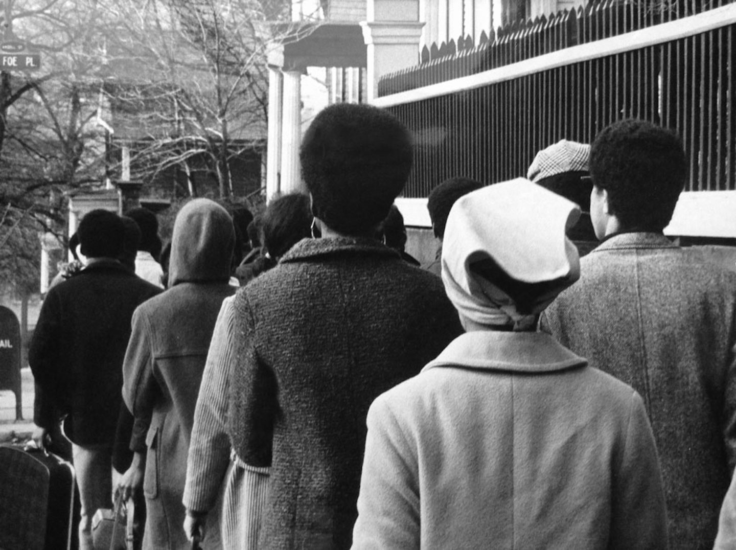 Black-and-white photo of a group of people walking in coats along a sidewalk, captured from behind, evoking a historic moment of community gathering or activism